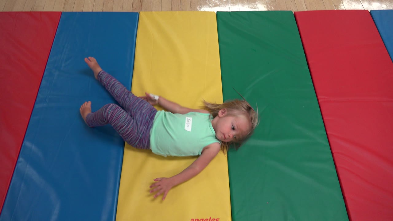 Young blonde girl in a mint top lies diagonally across blue, yellow, and green padded mats in Westminster, Colorado.