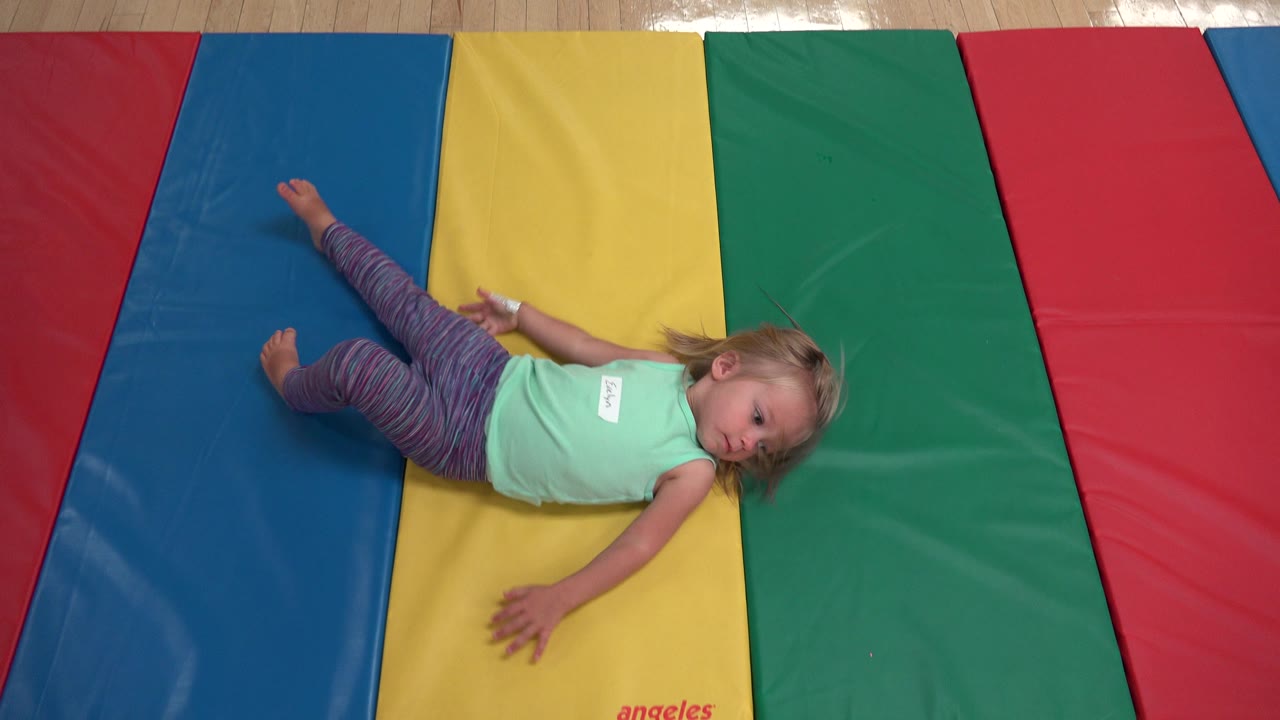 Young girl continues rolling with one arm extended over the yellow mat and her legs across the blue mat in Westminster, Colorado.