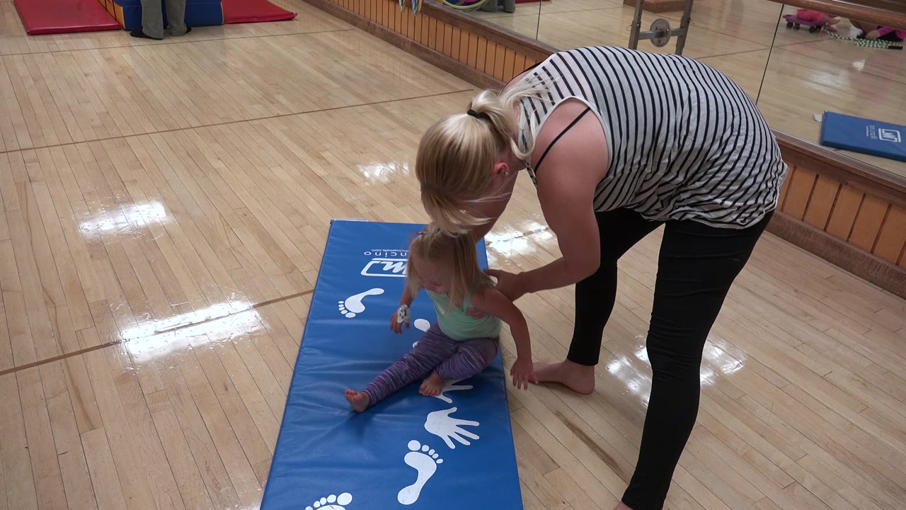 Woman bends over a young child sitting on a blue activity mat.