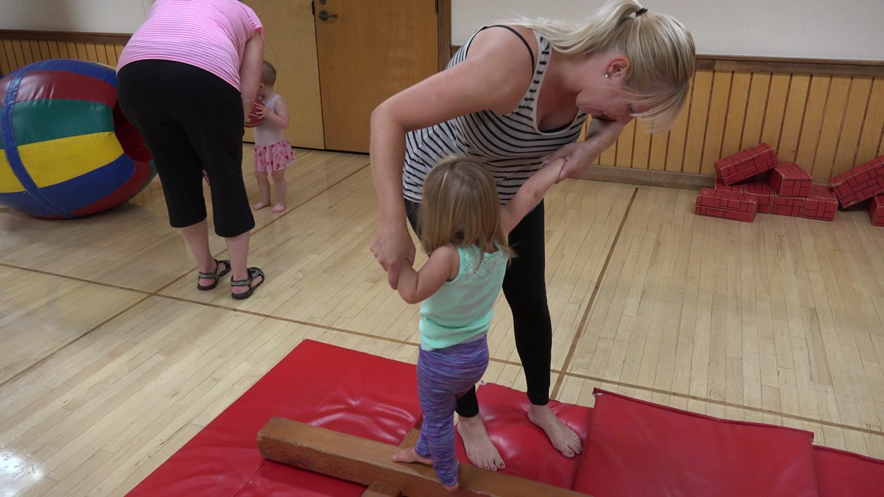 Adult woman guiding a small child on a balance beam in an indoor gym.