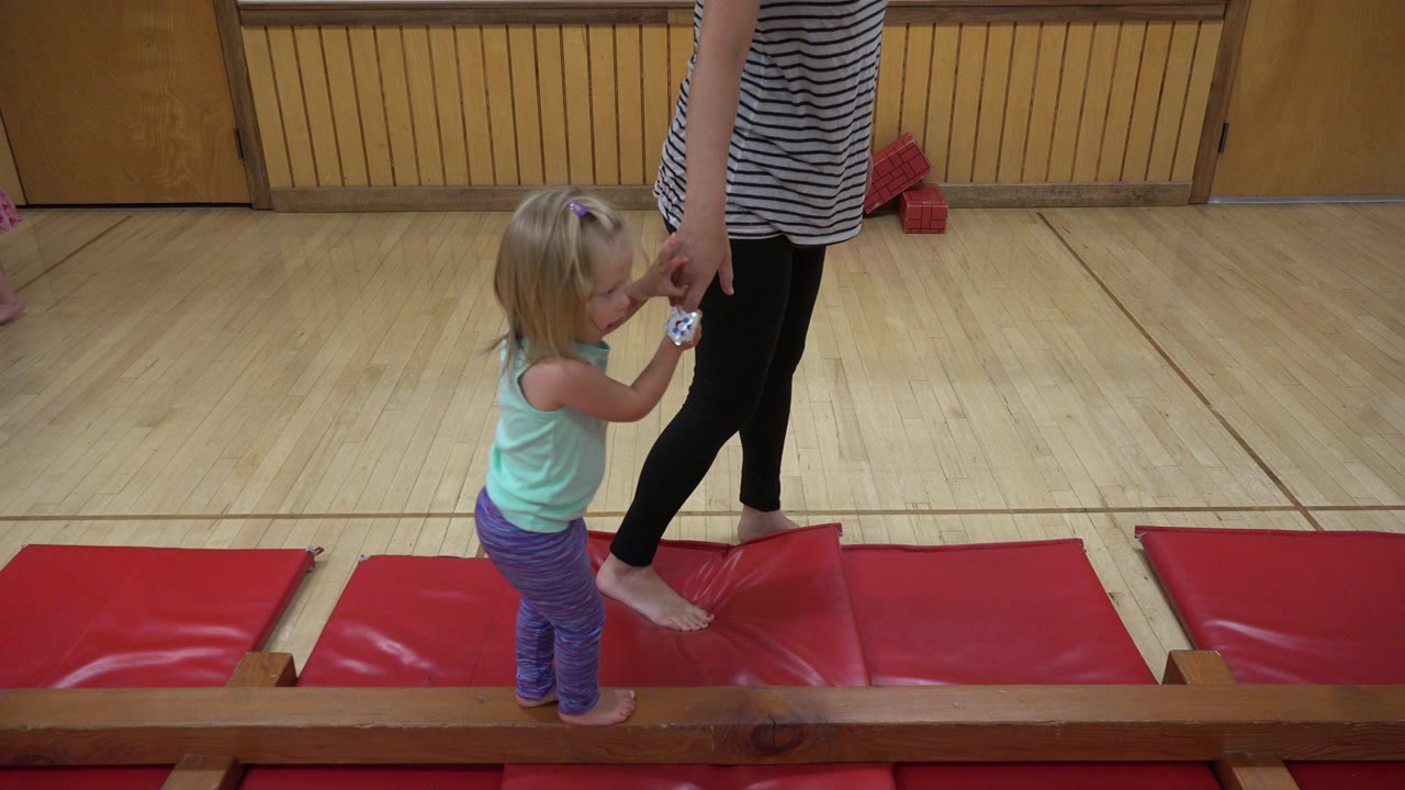 In Westminster, Colorado, the young girl looks down at her hand while the adult woman stands beside her on the wood floor near the red mats.