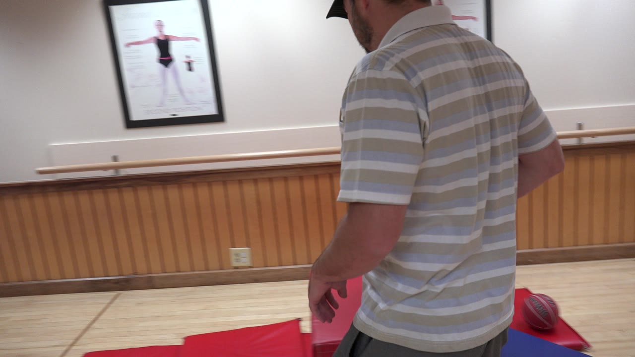 At the start of the Westminster, Colorado clip, a man in a striped shirt stands partly turned away near red mats, with a ballet barre and framed dance poster on the wall.
