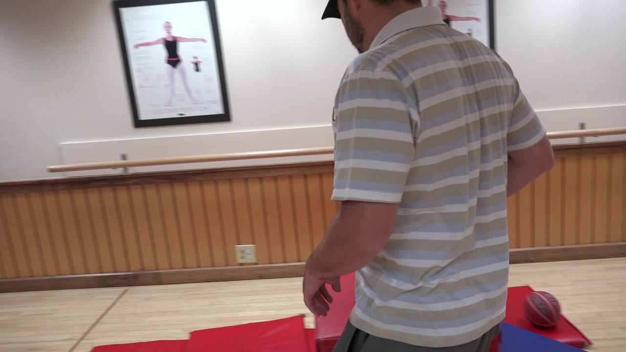 The man remains near the red mats in the Westminster, Colorado indoor room, with his back and shoulder closest to camera and dance instruction posters visible behind him.