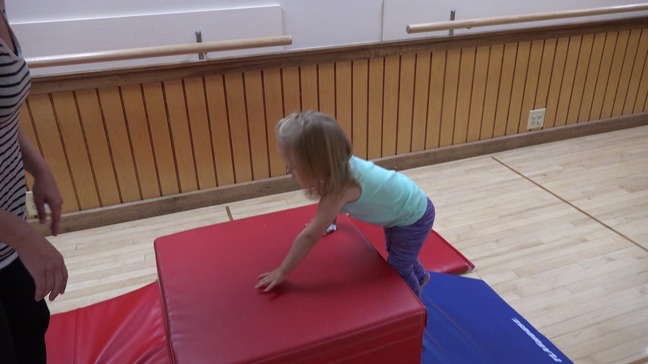 Child climbing onto a red padded gym block with an adult nearby.