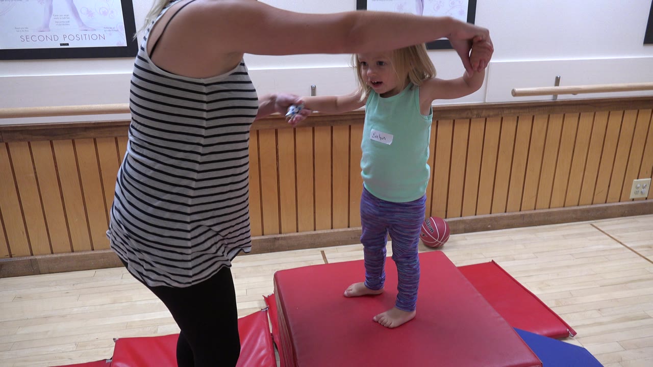 In Westminster, Colorado, a woman in a striped sleeveless top holds a young child's raised hand while the child stands barefoot on red mats near a wall barre.