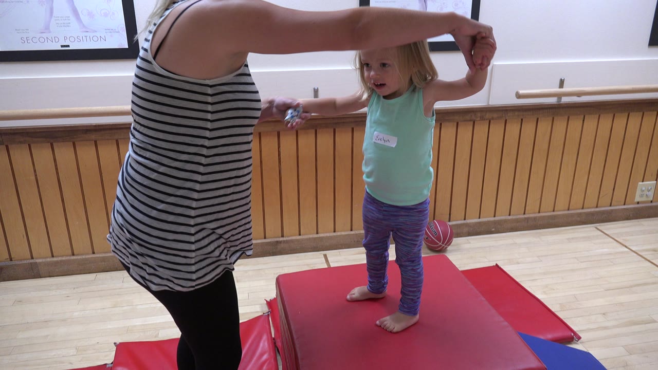 In Westminster, Colorado, the adult's arm stays extended above the child's head as the child faces forward on the red therapy mats beside the wood-paneled wall.
