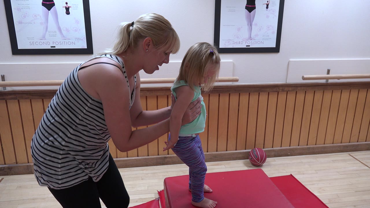 Adult woman steadies a young girl on a red gym mat.
