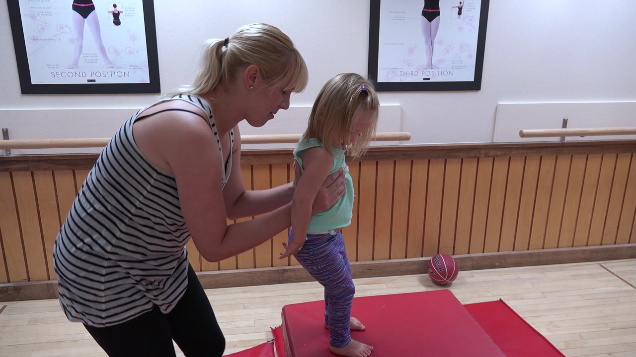 The adult woman keeps both hands near the young girl's shoulders as the child stands on the red mat in the Westminster indoor gym.