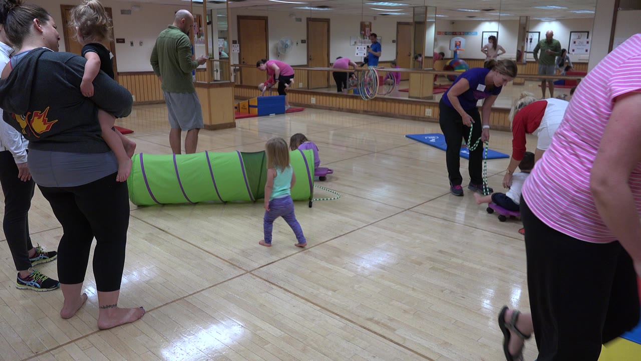 In Westminster, Colorado, adults stand near young children in a bright play room with a green tunnel, floor mats, hoops, and mirrored walls.