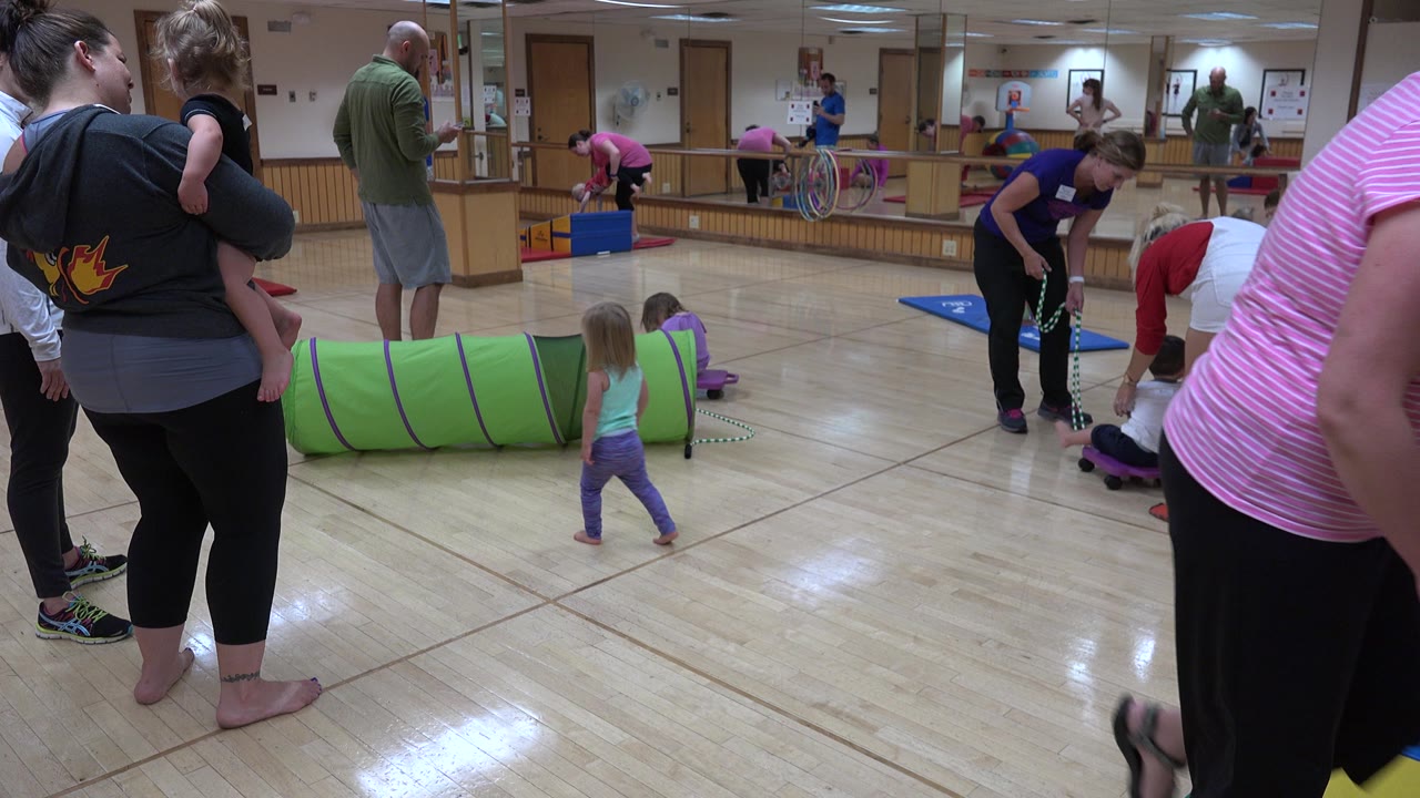 Adults and young children remain spread across the Westminster play room as a child stands near the green tunnel and toys on the wood floor.