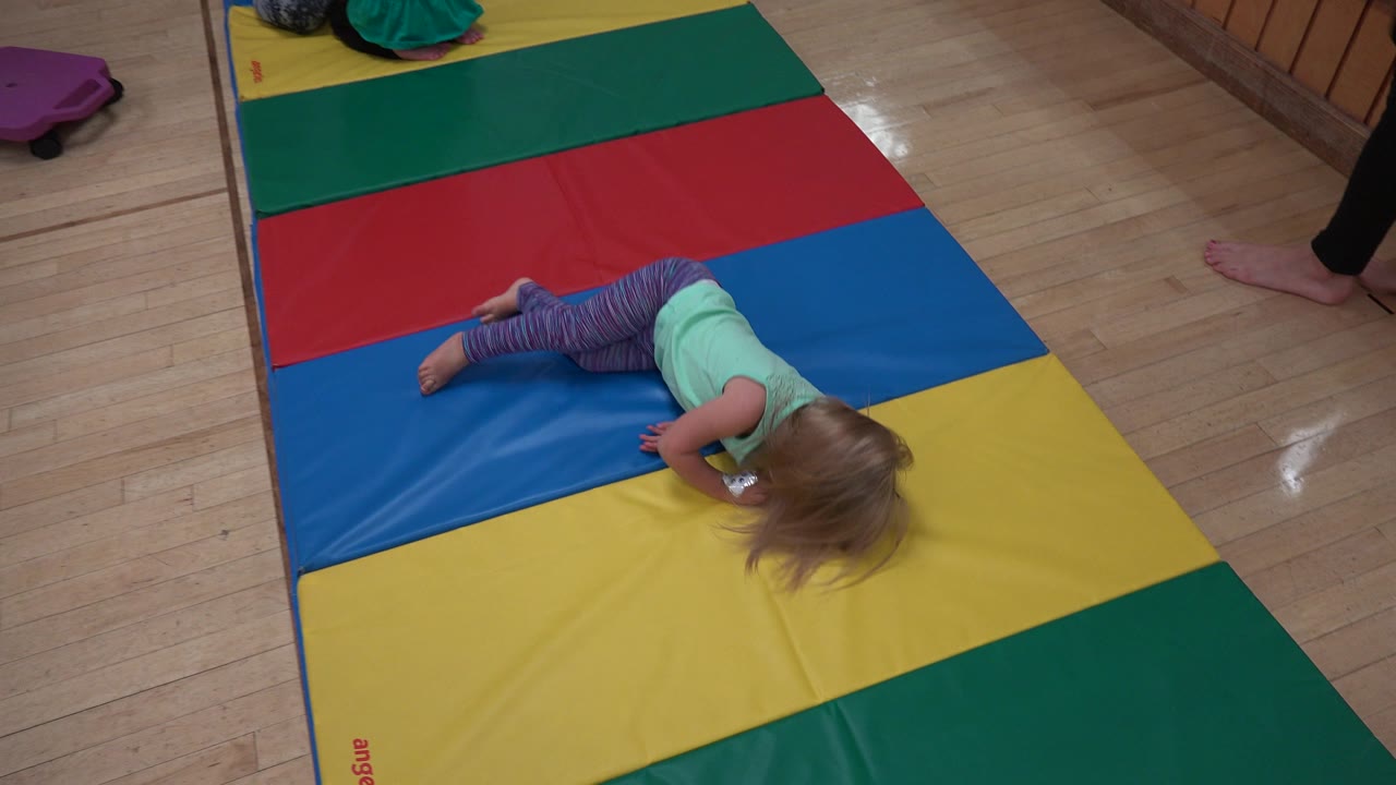 The child remains low on the yellow and blue mat panels in Westminster, Colorado, while a partial adult leg and another seated child appear near the top edge.