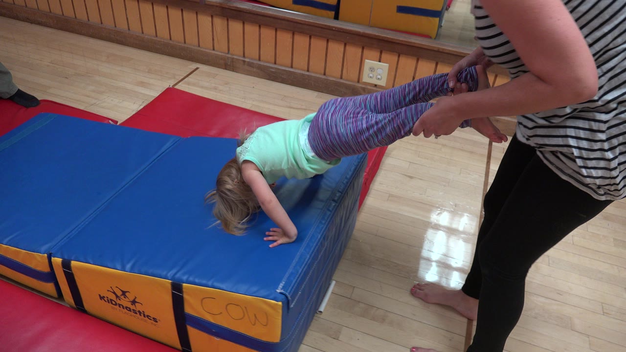 Adult holds a child by the legs as the child leans onto a blue gym mat.