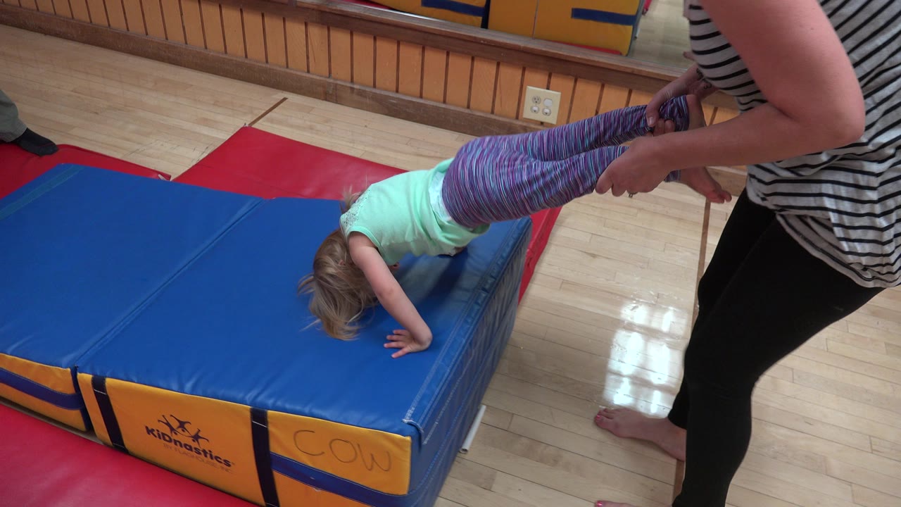 In Westminster, Colorado, the child remains bent over the blue padded mat as the adult steadies both legs near the mat edge during the therapy activity.