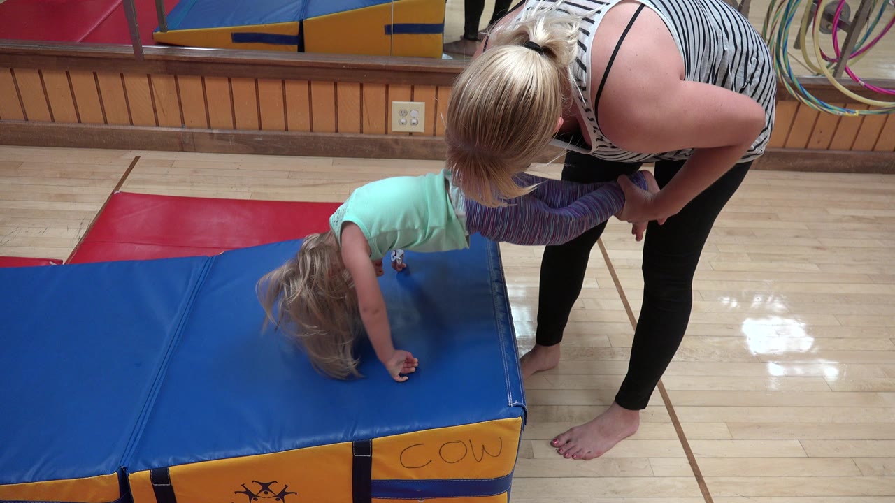 One second in, the child has both hands on the blue mat while the woman holds her legs and leans over to guide the movement in the Westminster play room.