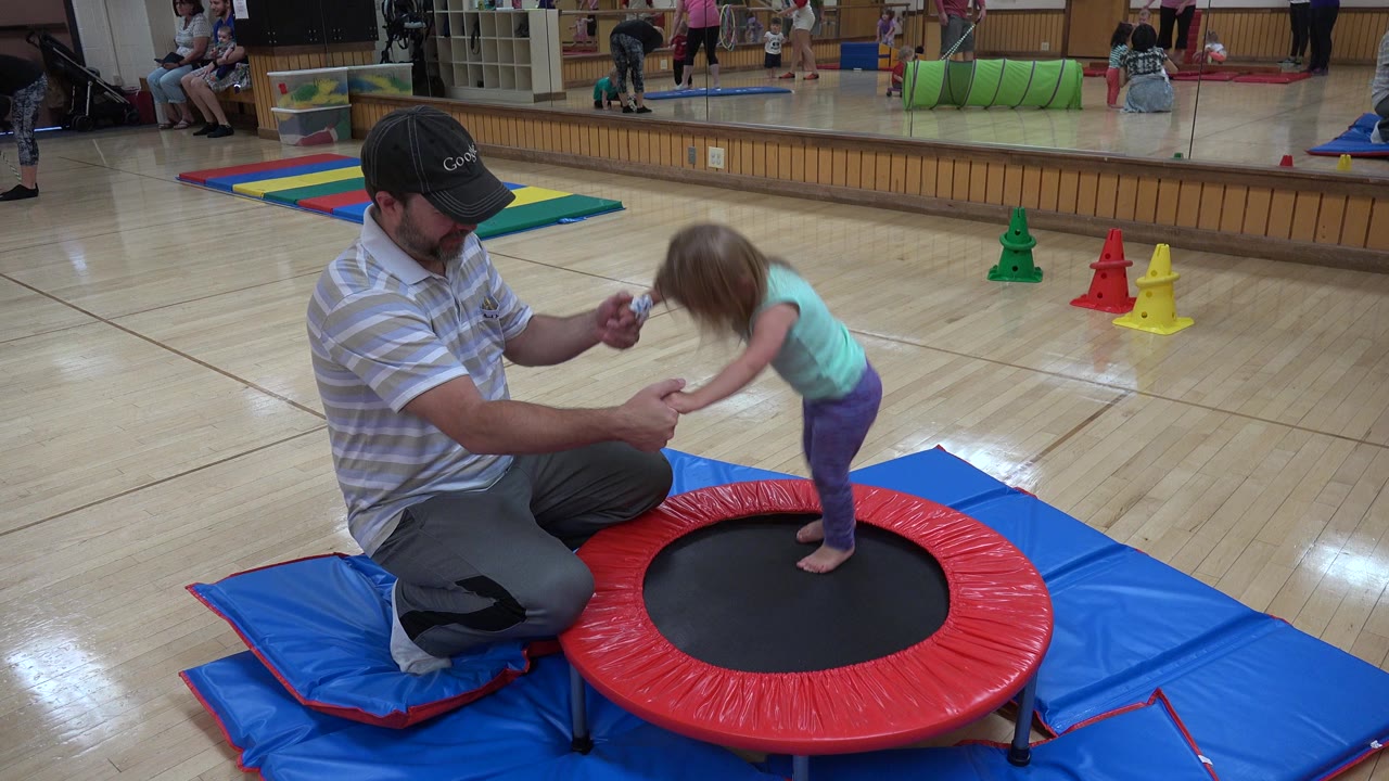 Man kneels beside a young girl on a small trampoline in an indoor activity room.
