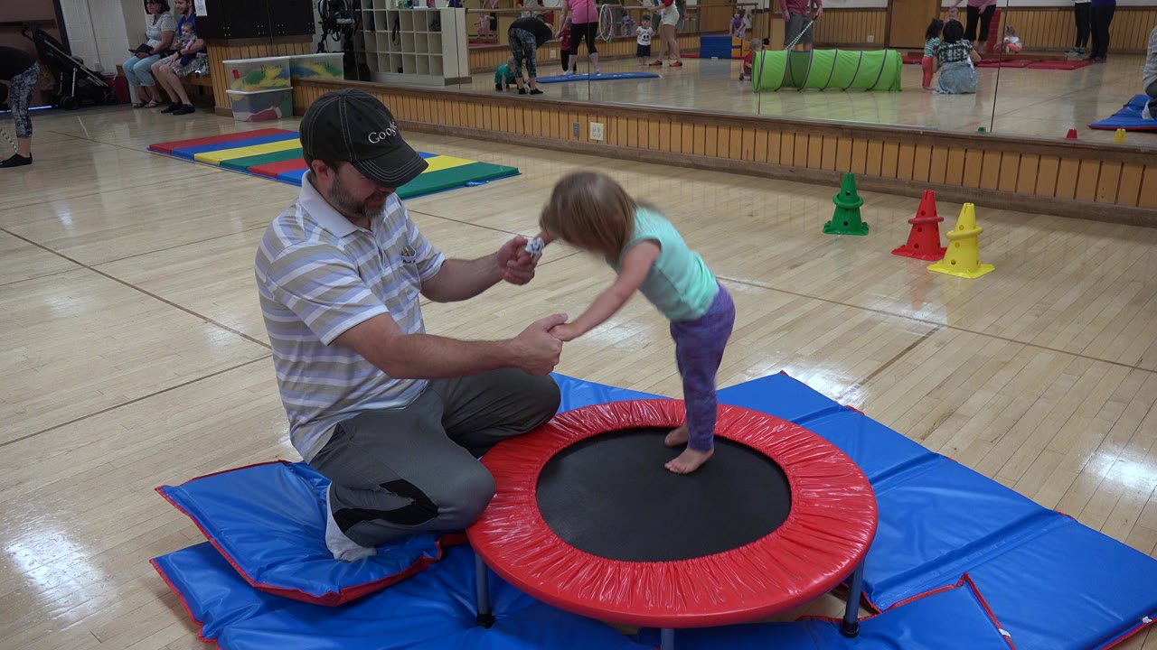 In Westminster, Colorado, the girl bends forward mid-bounce on the mini trampoline as the kneeling man keeps both hands near hers for support.