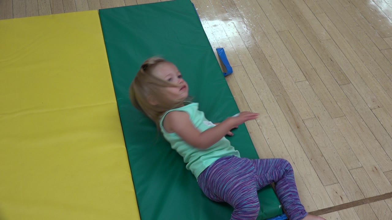 The child remains reclined on the green gym mat with one arm raised and the yellow mat running along her left side in Westminster, Colorado.