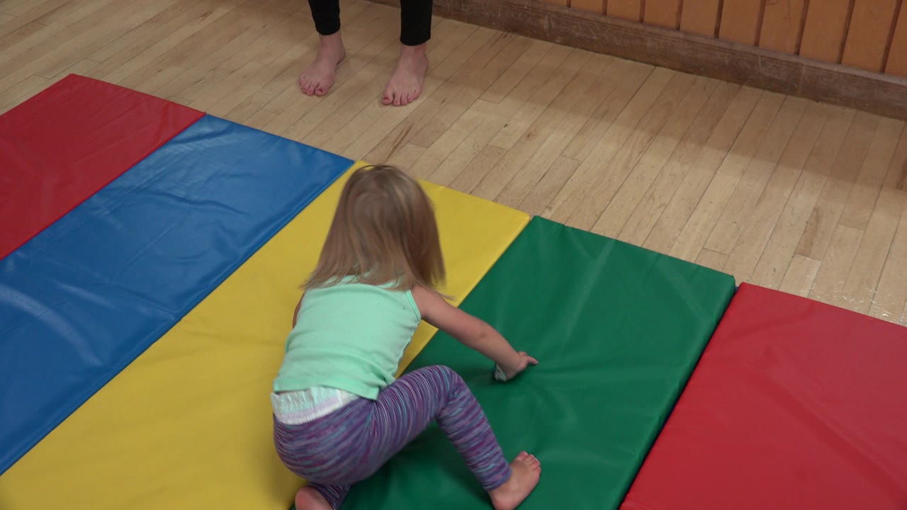A young blonde child crouches on green and yellow gym mats in Westminster, Colorado, with bare adult feet visible near the top of the frame.