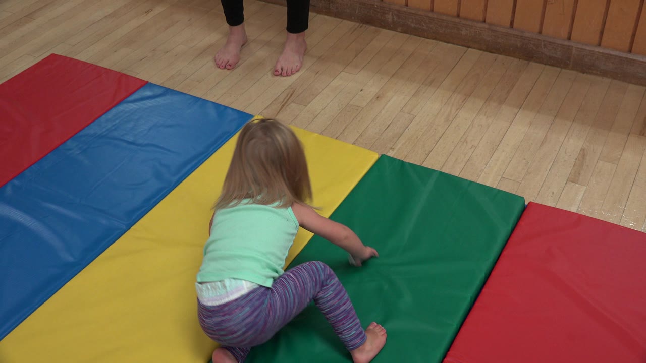 The child shifts forward on hands and knees across the yellow and green mats, with the polished wood floor and mirrored wall behind the play area.