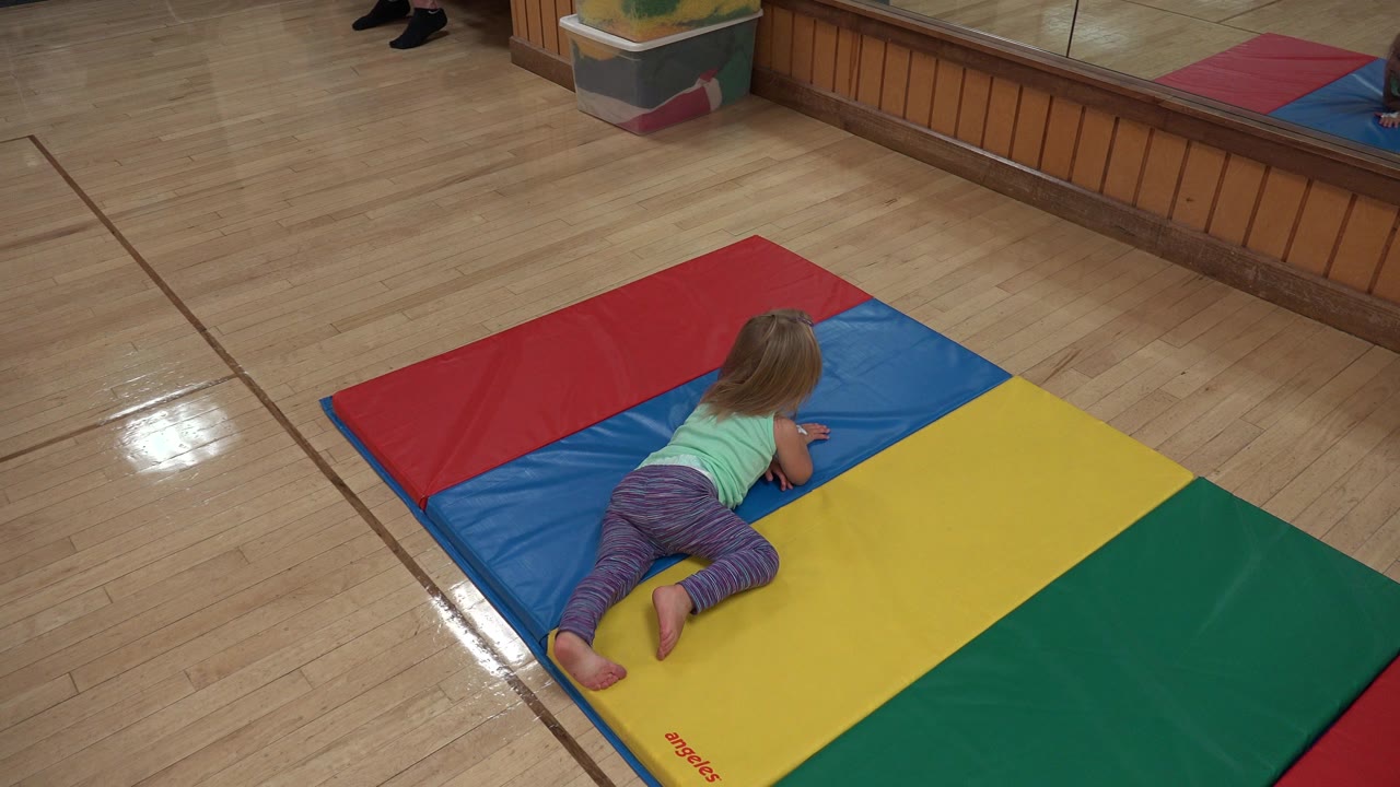 At 0:01, the girl remains on hands and knees near the blue and yellow panels of the padded mat, with the mirror and wood trim behind her.