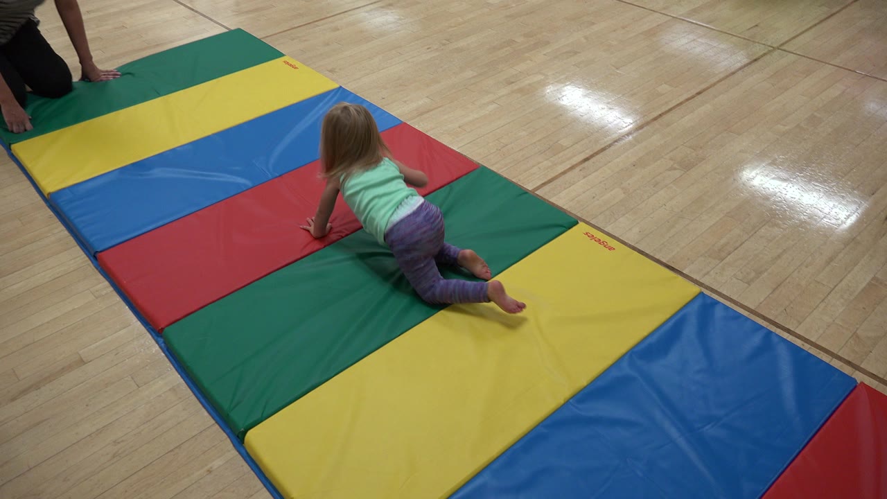 Baby crawling on colorful floor mats with an adult partly visible nearby.