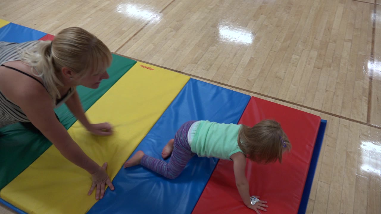 Adult woman watches a young girl crawling on colorful gym mats.