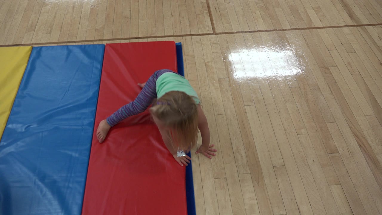 The child remains low to the red mat as she crawls forward, with blue and yellow padded sections along the left side and gym floor visible beyond.