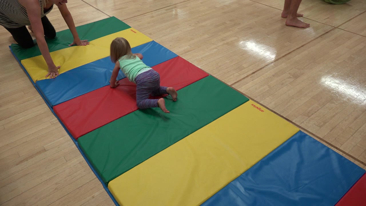 Young girl crawls on a colorful padded mat while an adult kneels nearby.