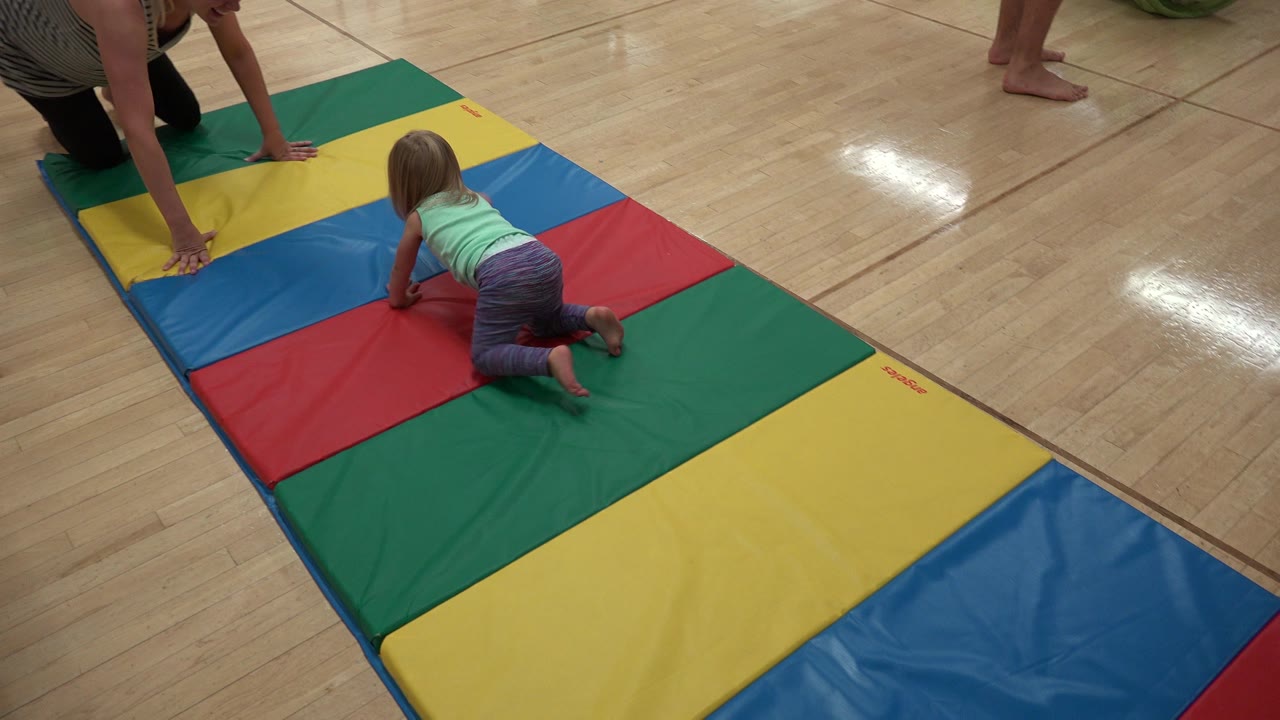 At 0:01 in Westminster, Colorado, the child has moved forward on the red and blue mat panels while the kneeling adult remains near the top of the mat.