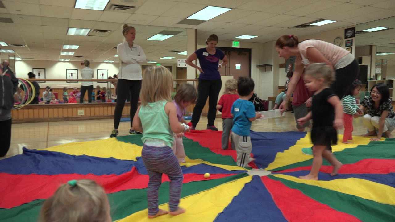 Children stand and move across the multicolor activity mat in the Westminster indoor gym, with a woman in white and other adults visible behind them.