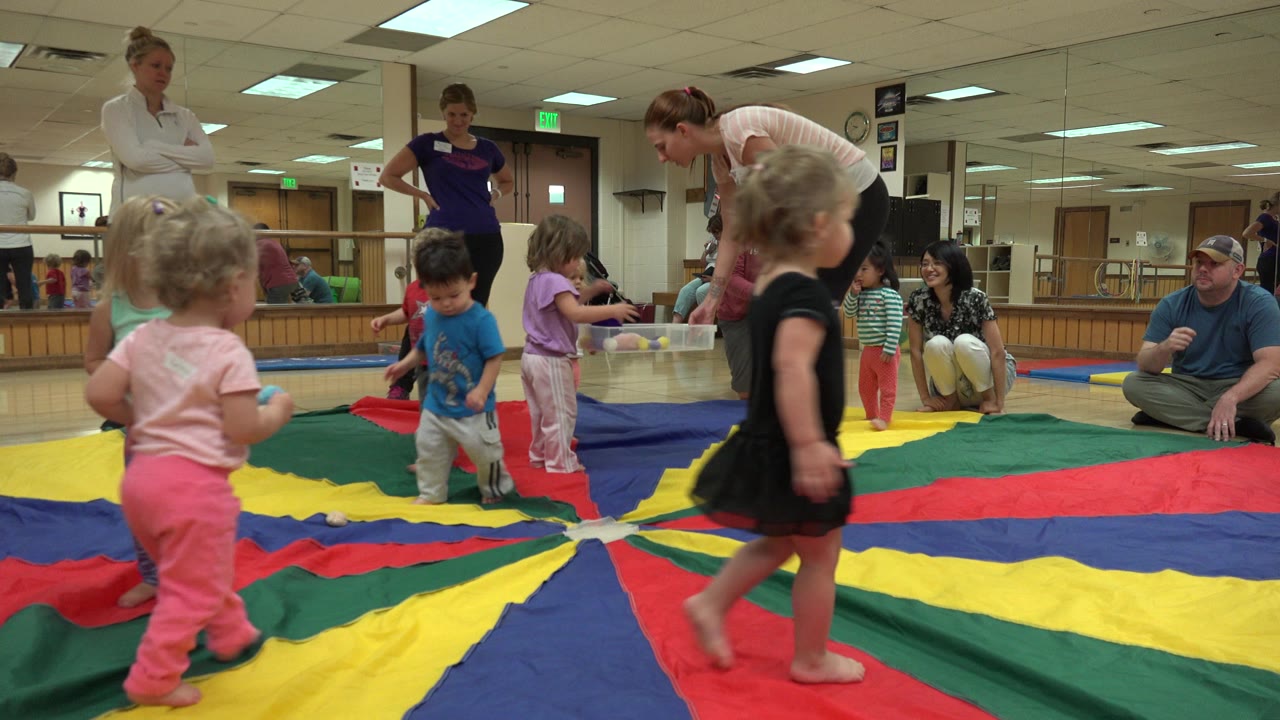 Toddlers and women around a colorful parachute mat in an indoor playroom.
