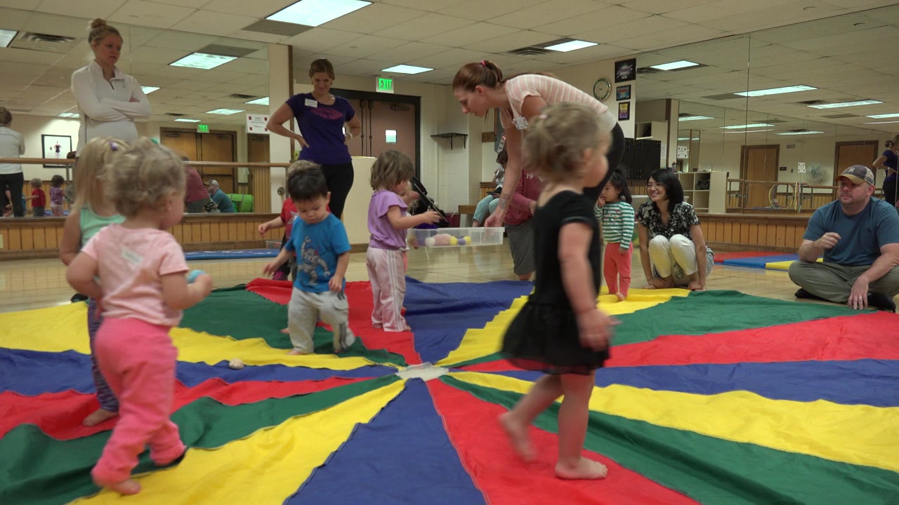 Children cluster near the center of a colorful play parachute as two standing women and seated adults watch inside the Westminster playroom.