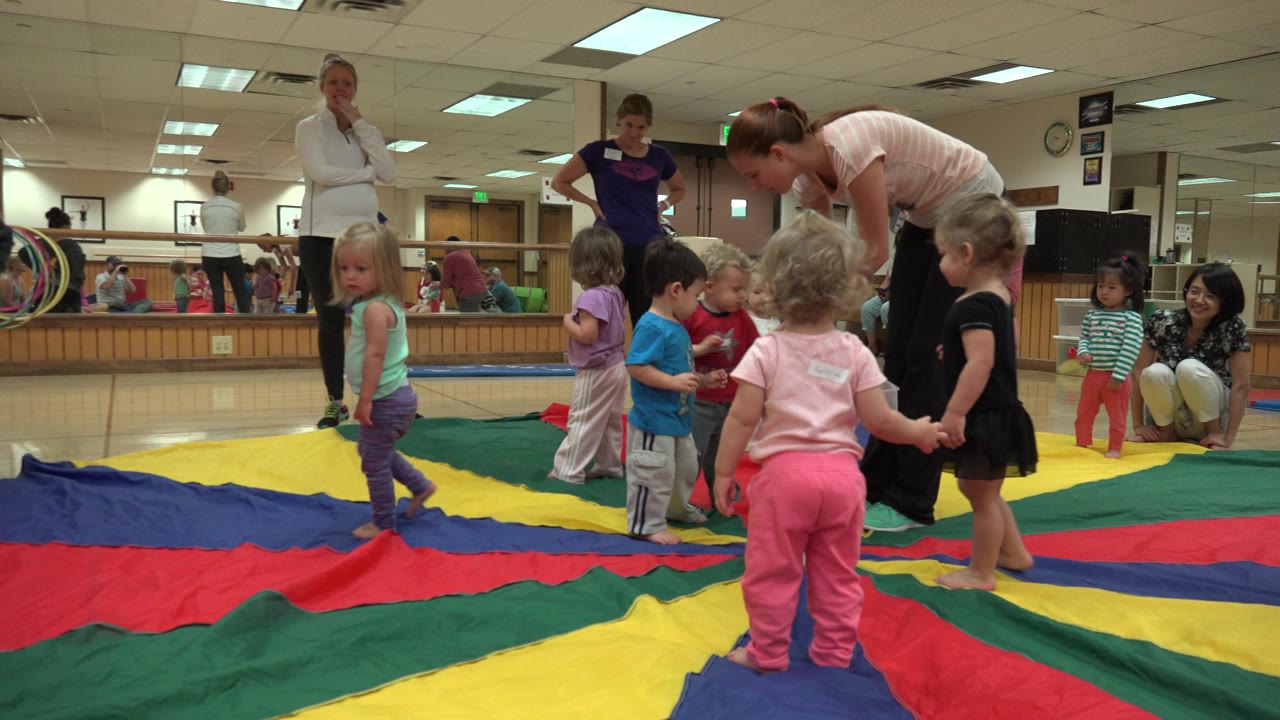 Toddlers stand on a red, yellow, green, and blue parachute mat while adults supervise in an indoor Westminster, Colorado play room with mirrors along the wall.