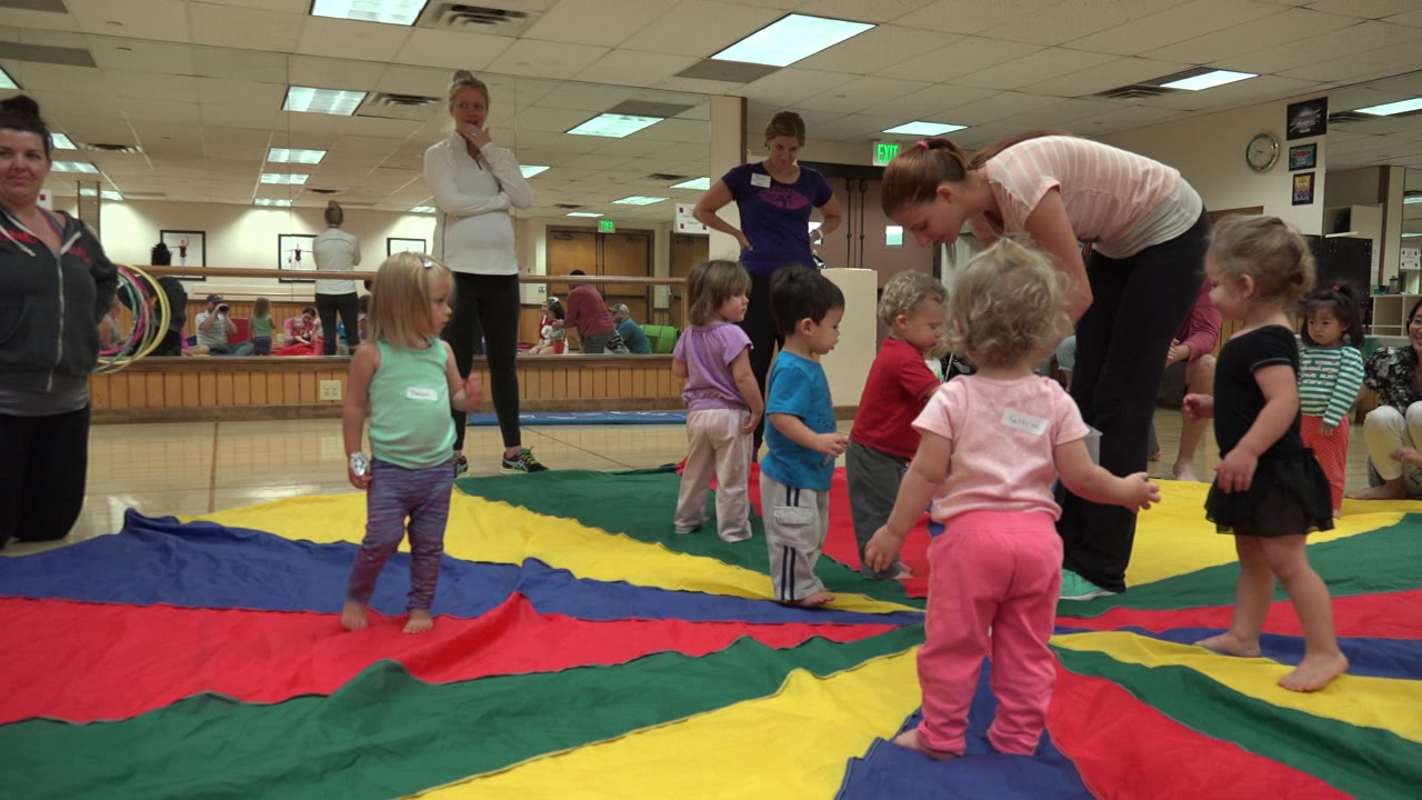 A kneeling adult and toddlers on a colorful mat in an indoor play room.