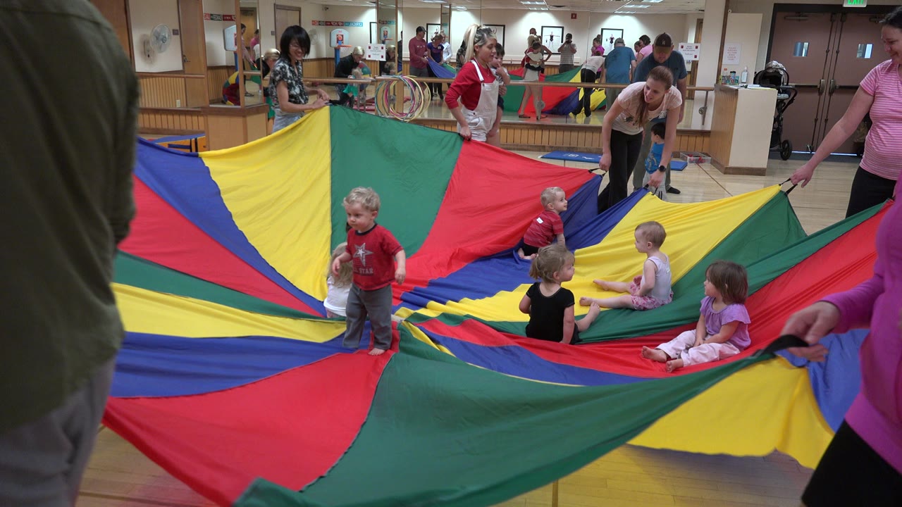 Parents and children around a colorful play parachute in an indoor room.
