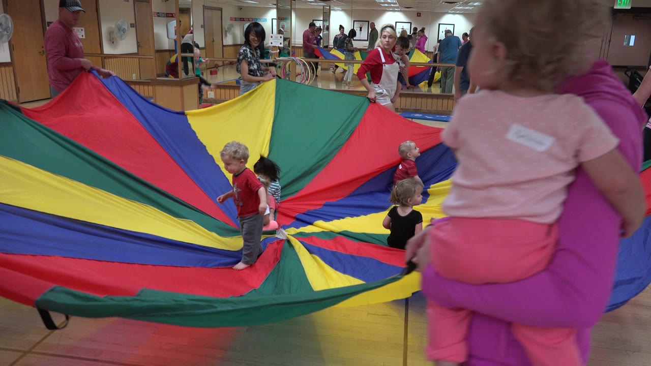 Adult carrying a toddler near a colorful parachute activity.