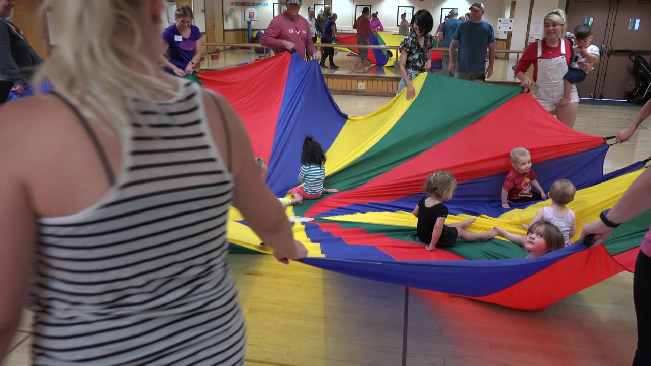 In Westminster, Colorado, adults stand around a red, yellow, green, and blue play parachute as young children sit and look around inside the indoor activity circle.