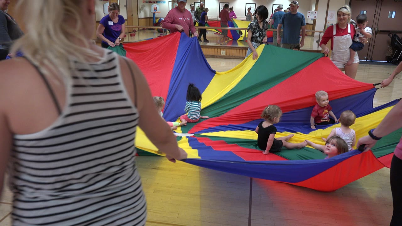 In Westminster, Colorado, the parachute lifts slightly around seated children while adults in casual clothes hold the edge inside a wood-floor room.