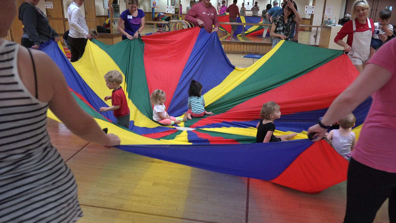 Adults pull the colorful parachute outward with several toddlers near the center.