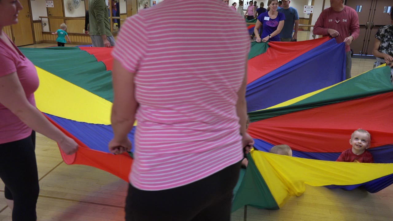 Adults hold a multicolored parachute while a toddler looks up from inside the fabric.