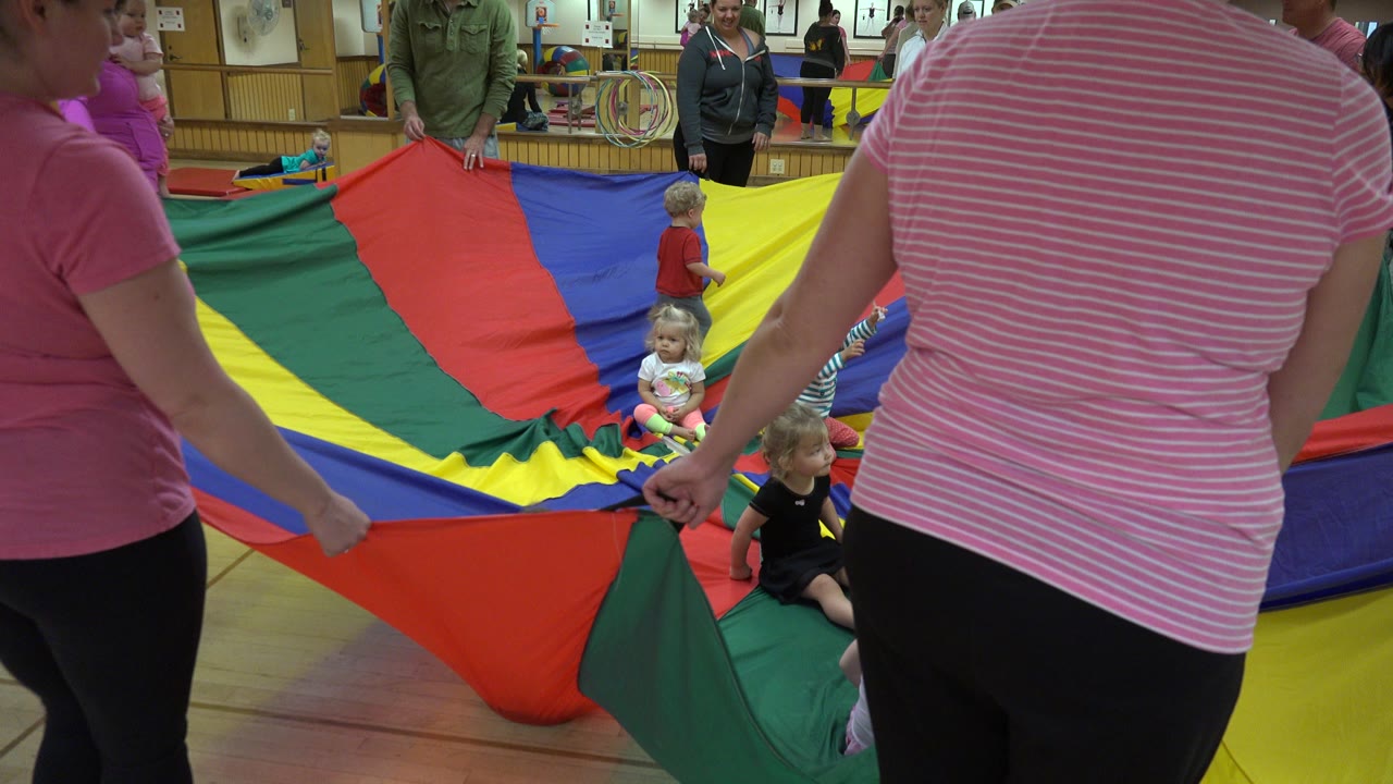 Adults hold the edges of a rainbow parachute while young children sit in the center during a Westminster, Colorado activity.