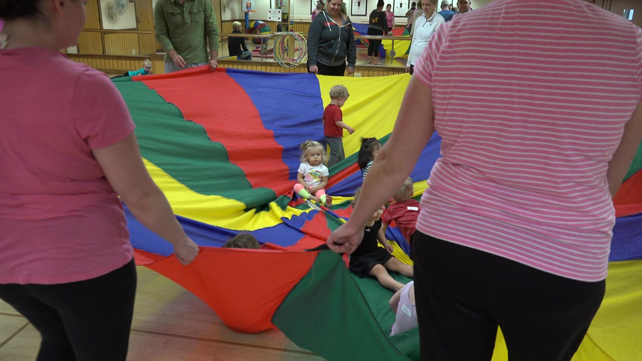 Adults stand around the parachute with children clustered near the center.