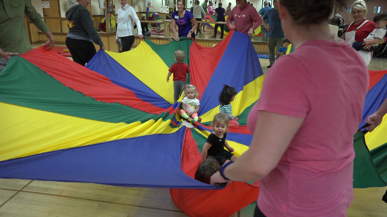 Adults and children around a colorful play parachute indoors.