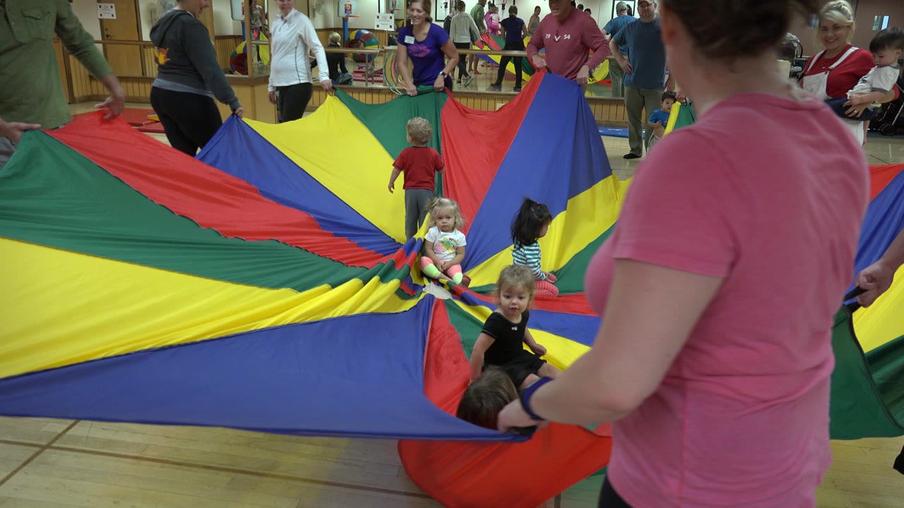 A toddler faces the camera near the front of the parachute while adults around the perimeter hold the stretched fabric in the Westminster indoor activity space.