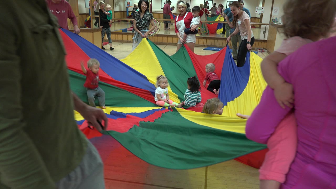 Adults and toddlers around a colorful parachute sheet indoors.