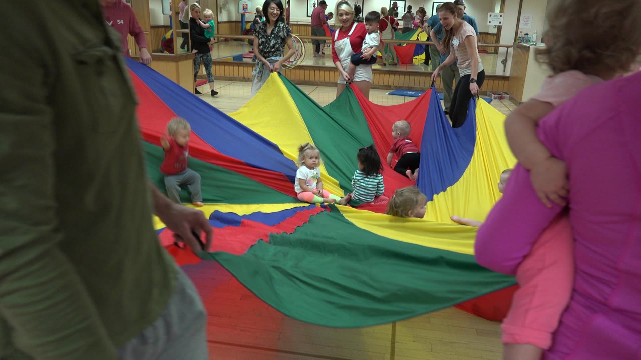 In Westminster, Colorado, several adults lift the edges of a bright parachute sheet as small children sit and kneel on the fabric in the middle of the room.