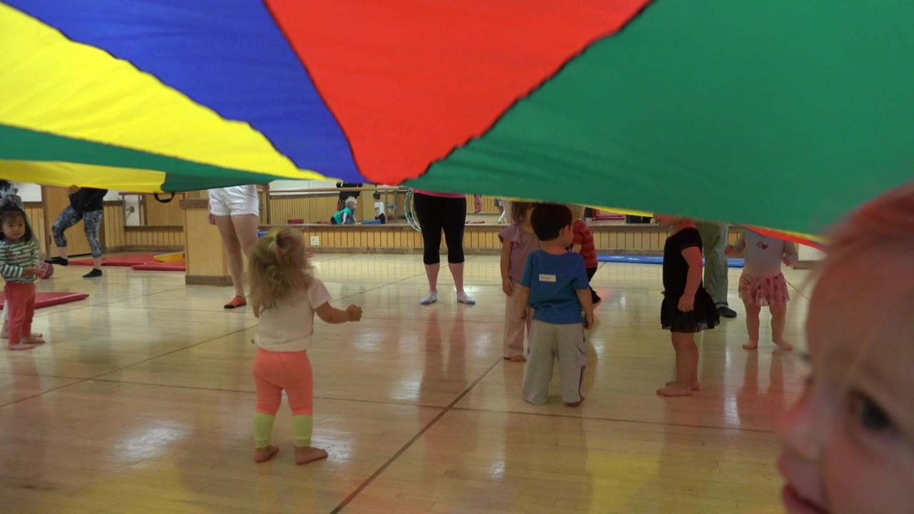 Children and adults under a multicolored play sheet in an indoor room.