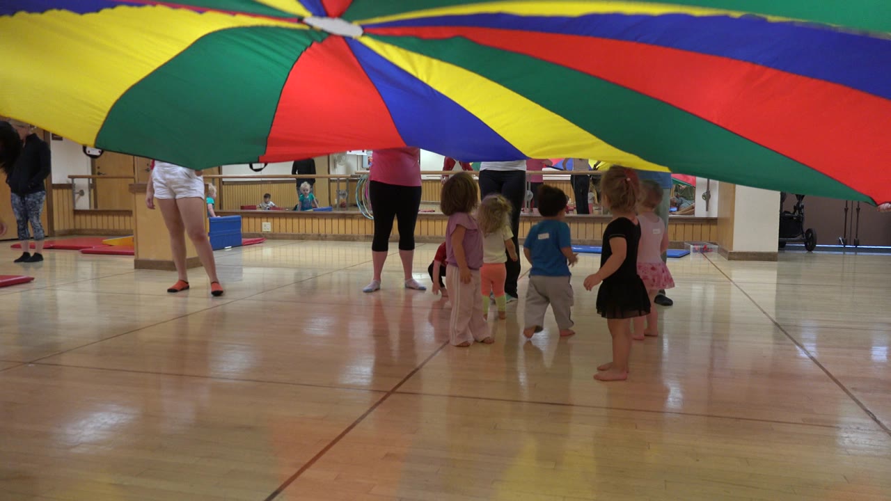 Children and adults under a colorful play parachute in an indoor studio.