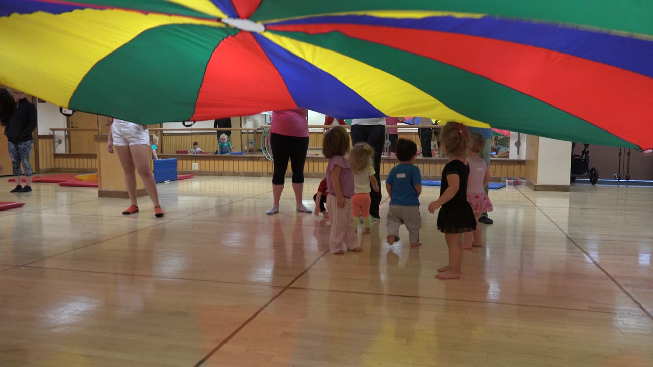 In Westminster, Colorado, the multicolor parachute remains overhead as children stand near adults on the polished floor of a mirrored activity room.