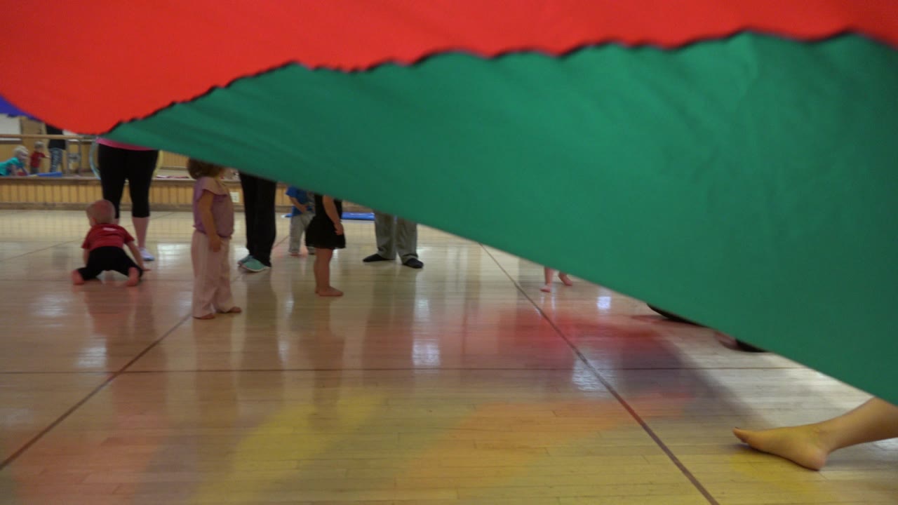 One second in, the colorful parachute still fills the upper frame while children and adults stand or crawl beneath it in the Westminster indoor play space.